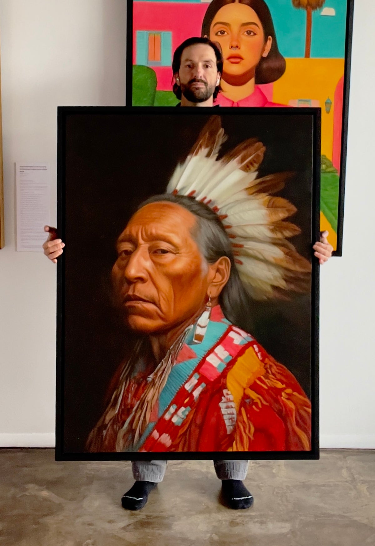 MEFREEK Seth Jennemann Original Oil Painting hyperrealistic portrait of a Native American man wearing a traditional feathered headdress with detailed skin texture and solemn expression dressed in richly colored ceremonial garments with intricate beadwork and fringe set against a deep dark background with dramatic lighting and refined realism while the painting is being held in a gallery space revealing the artist partially visible behind the canvas with a contemporary colorful portrait hanging in the backgr